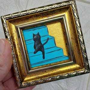 May include: A black cat painted on a blue and gold background, framed in a gold ornate frame.