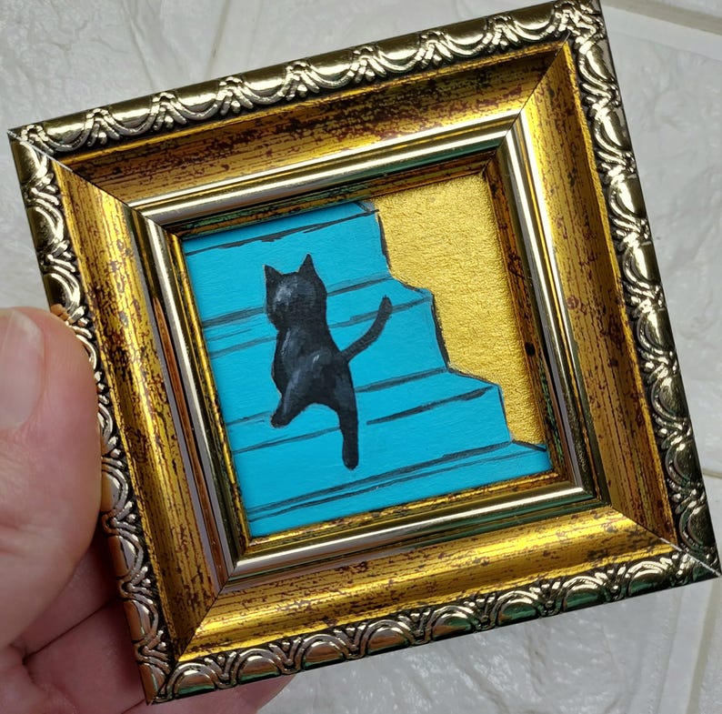 May include: A black cat painted on a blue and gold background, framed in a gold ornate frame.