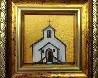 May include: A small white church with a black door and windows, painted on a gold background. The church is framed in a gold frame.