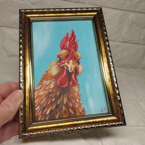 Puede incluir: Pintura enmarcada de la cabeza de un gallo. El gallo tiene plumas naranjas y marrones, una cresta y barbilla rojas, sobre un fondo azul claro. El marco es dorado con detalles ornamentados.