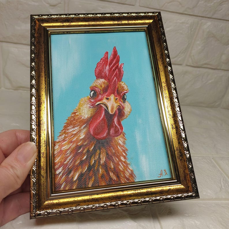 Puede incluir: Pintura enmarcada de la cabeza de un gallo. El gallo tiene plumas naranjas y marrones, una cresta y barbilla rojas, sobre un fondo azul claro. El marco es dorado con detalles ornamentados.