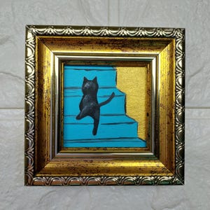 May include: A framed painting of a black cat walking up a set of blue stairs. The frame is gold with a silver trim.