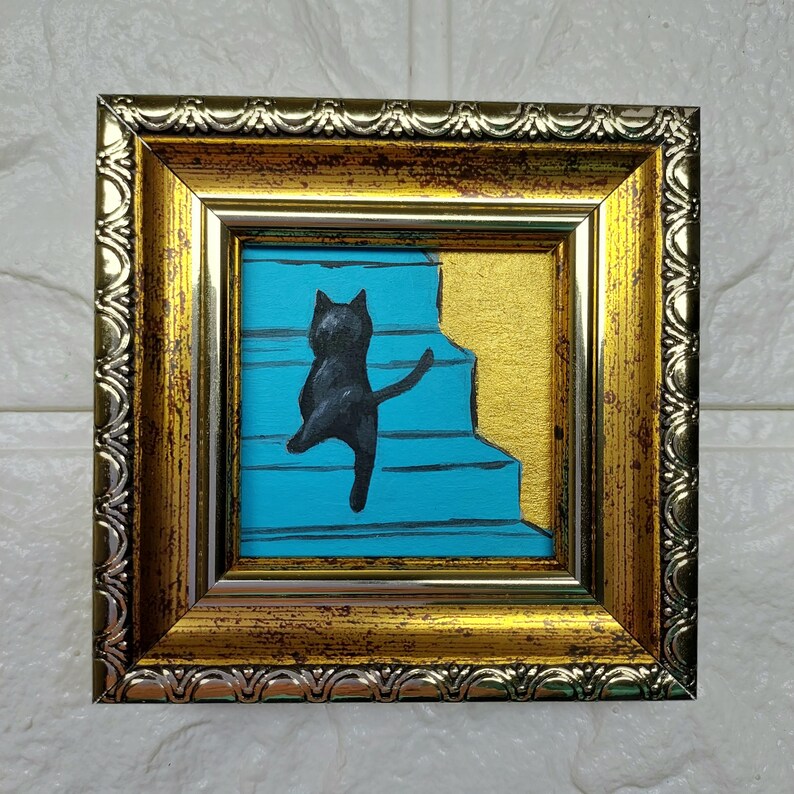 May include: A framed painting of a black cat walking up a set of blue stairs. The frame is gold with a silver trim.