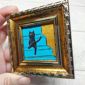 May include: A black cat painted on a blue and gold background, climbing a set of stairs. The painting is framed in a gold frame.