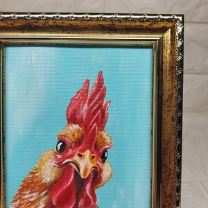 Puede incluir: Pintura enmarcada de un gallo con cresta y barbilla rojas, sobre un fondo azul claro. El plumaje del gallo es una mezcla de amarillo, naranja y marr&oacute;n. El marco tiene un acabado dorado ornamentado.