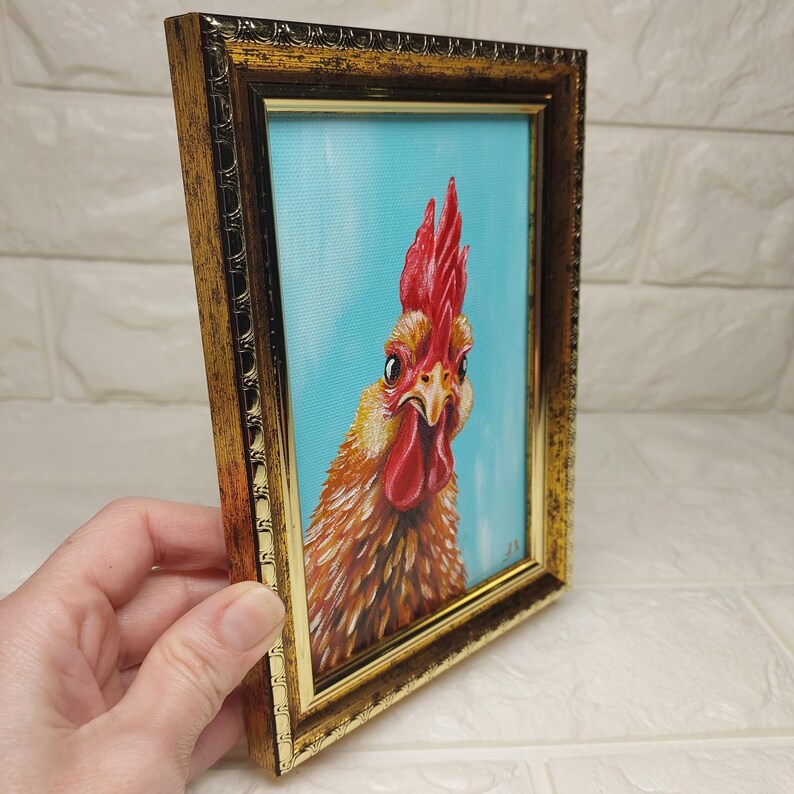 Puede incluir: Pintura enmarcada de un gallo con cresta y barbilla rojas, sobre un fondo azul claro. Las plumas del gallo son de tonos marrones y dorados. El marco es ornamentado con un acabado dorado.