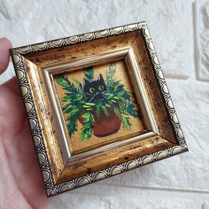 May include: A black cat peeking out of a potted plant, painted in a miniature style and framed in a gold and silver frame.