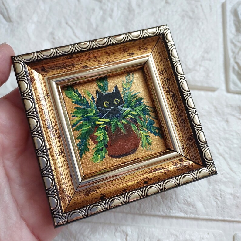 May include: A black cat peeking out of a potted plant, painted in a miniature style and framed in a gold and silver frame.