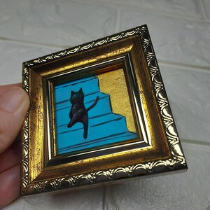 May include: A black cat painted on a blue and gold background, climbing a set of stairs. The painting is framed in a gold frame with a decorative border.