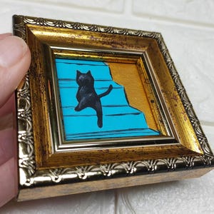 May include: A black cat painted on a blue background with gold accents, climbing a set of stairs. The painting is framed in a gold frame with a decorative border.
