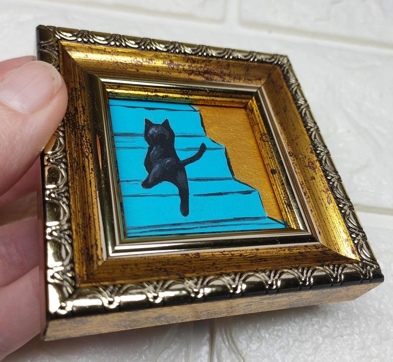 May include: A black cat painted on a blue background with gold accents, climbing a set of stairs. The painting is framed in a gold frame with a decorative border.