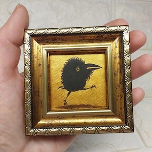 以下が含まれることがあります： 明るい金色の背景に描かれた、黄色い目を持つ黒い鳥の小さな正方形の絵画。この作品は、質感のあるスカラップ状の縁とダークブラウンのアンティーク調のディテールが施された、装飾的な金色のフレームに収められています。