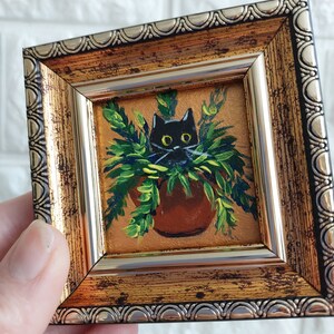 May include: A black cat peeking out from a green plant in a terracotta pot. The painting is framed in a gold and silver ornate frame.