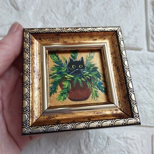 May include: A small framed painting of a black cat peeking out of a green plant pot. The frame is gold with a silver trim.
