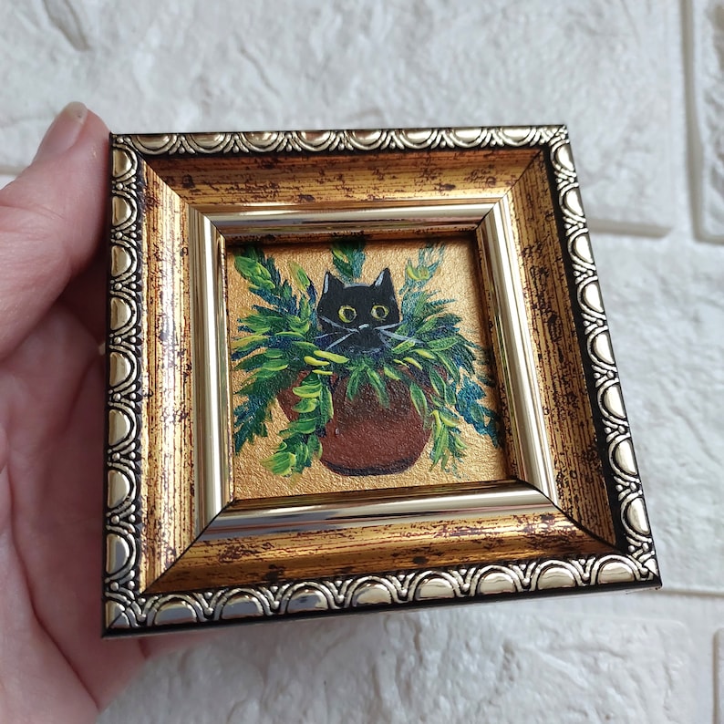 May include: A small framed painting of a black cat peeking out of a green plant pot. The frame is gold with a silver trim.