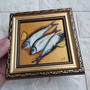 May include: A small square painting in a dark brown and gold ornate frame featuring three silver fish arranged diagonally on a textured gold background with two yellow lemon wedges. The artist's initials A3 are visible in the bottom right corner.