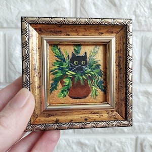 May include: A black cat peeking out of a potted plant, painted in a miniature style. The painting is framed in a gold and silver ornate frame.
