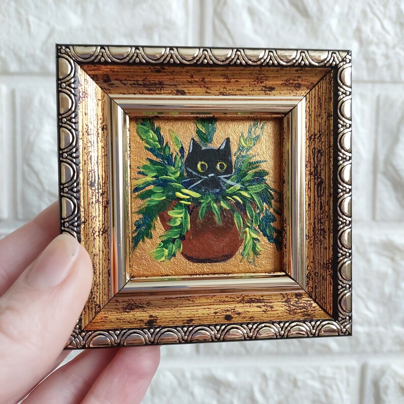 May include: A black cat peeking out of a potted plant, painted in a miniature style. The painting is framed in a gold and silver ornate frame.