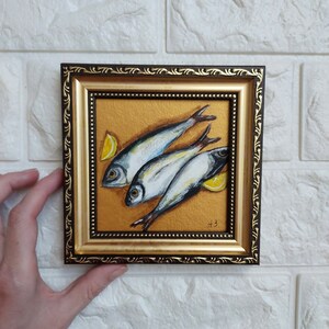 May include: A square painting in a dark brown and gold ornate frame featuring three silver fish arranged diagonally on a textured gold background with two yellow lemon wedges. The artist's initials A.B. are visible in the bottom right corner.