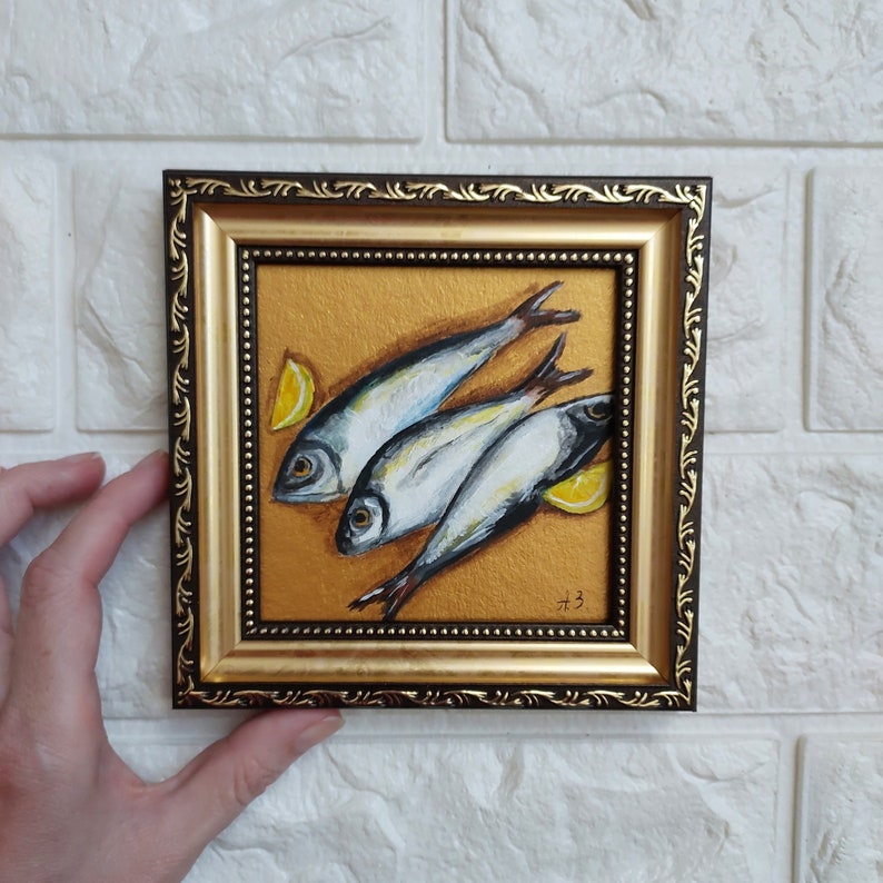 May include: A square painting in a dark brown and gold ornate frame featuring three silver fish arranged diagonally on a textured gold background with two yellow lemon wedges. The artist's initials A.B. are visible in the bottom right corner.