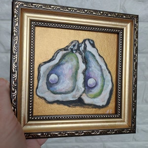 May include: A square painting of two open oysters with iridescent green and purple interiors, each containing a white pearl. The artwork is set against a metallic gold background and enclosed in a decorative dark brown and gold ornate frame.