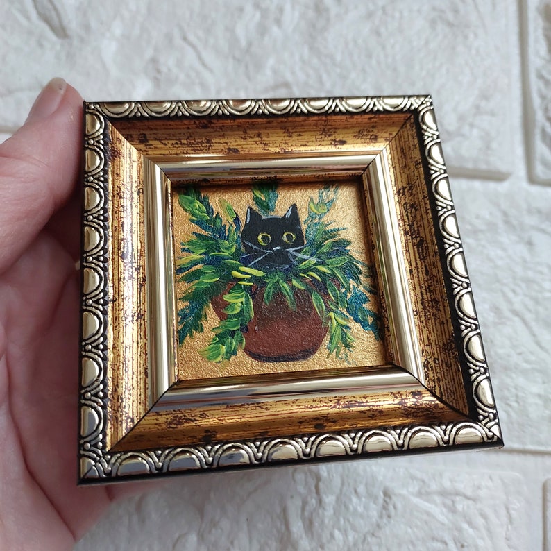 May include: A small framed painting of a black cat peeking out of a green plant pot. The frame is gold with a silver trim.