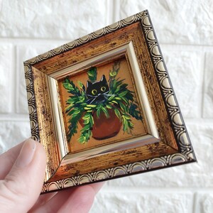 May include: A black cat with yellow eyes peeks out from a pot of green plants. The painting is framed in a gold ornate frame.