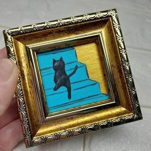 May include: A black cat silhouette climbing a blue staircase, framed in a gold ornate frame.