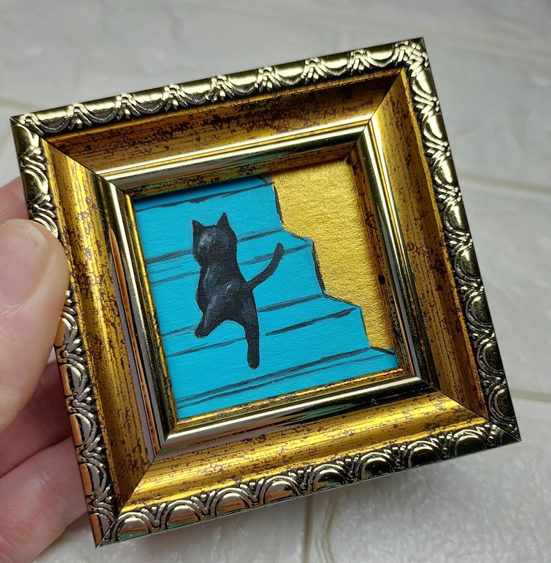 May include: A black cat silhouette climbing a blue staircase, framed in a gold ornate frame.