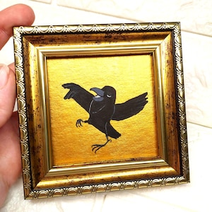 May include: A small square painting featuring a black crow with its wings spread wide, set against a vibrant metallic gold background. The artwork is encased in an ornate, antique-style gold frame with intricate embossed detailing along the edges.