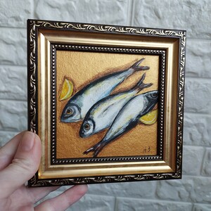 May include: A square framed painting featuring three silver fish arranged diagonally on a textured metallic gold background, accompanied by two yellow lemon wedges. The frame is dark brown with ornate gold detailing and a beaded inner border.