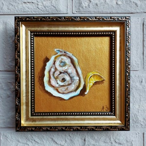 Könnte beinhalten: Ein gerahmtes Gemälde einer Auster mit einem Zitronenspalten auf goldenem Hintergrund. Das Gemälde ist unten rechts mit "A3" signiert.