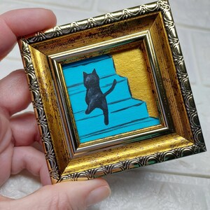 May include: A black cat silhouette painted on a blue and gold background, framed in a gold ornate frame.