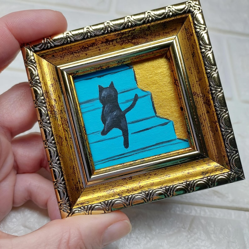May include: A black cat silhouette painted on a blue and gold background, framed in a gold ornate frame.