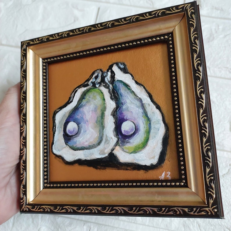 May include: A square framed painting featuring two open oyster shells with iridescent purple and green interiors, each containing a white pearl. The artwork is set against a solid gold background and enclosed in a dark brown and gold ornate frame.