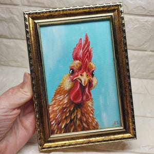 Puede incluir: Pintura enmarcada de un gallo con cresta y barbilla rojas, sobre un fondo azul claro. Las plumas del gallo son de tonos naranja y marr&oacute;n. El marco es dorado con detalles ornamentados.
