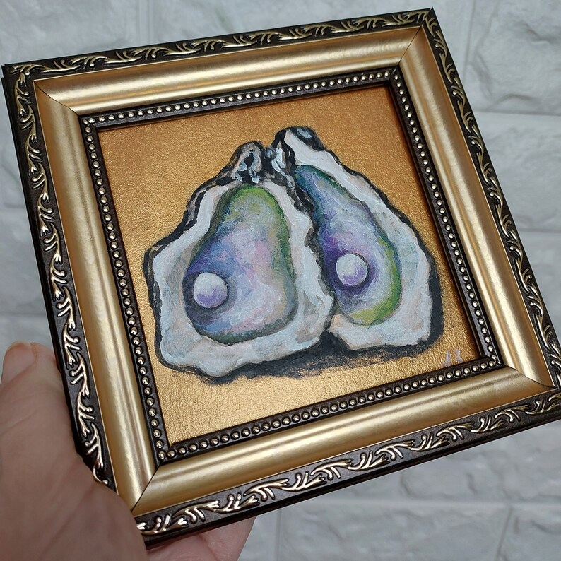 May include: A square painting of two open oyster shells with iridescent interiors and white pearls, set against a metallic gold background. The artwork is enclosed in a decorative, ornate gold and dark brown frame with a beaded inner edge detail.