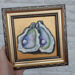 May include: A square framed painting featuring two open oyster shells with iridescent interiors and a single white pearl in each, set against a solid gold background. The frame is dark brown with gold accents and a beaded inner edge detail.