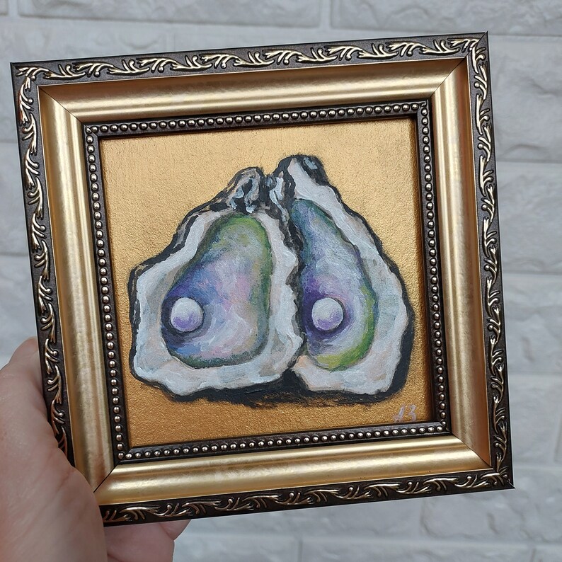 May include: A square framed painting featuring two open oyster shells with iridescent interiors and a single white pearl in each, set against a solid gold background. The frame is dark brown with gold accents and a beaded inner edge detail.