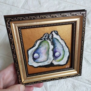 May include: A square painting of two open oysters with iridescent purple, green, and white interiors, set against a metallic gold background. The artwork is enclosed in a dark brown and gold ornate frame with a beaded inner edge and initials AB.