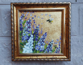 May include: A square painting in a decorative gold frame featuring a cluster of purple and white flowers against a textured gold background. A small bee is depicted in flight near the top right, with scattered petals floating in the air around it.