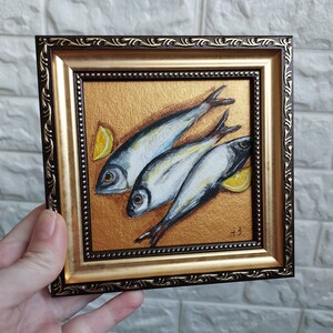 May include: A square painting in a dark brown and gold ornate frame featuring three silver fish arranged diagonally on a textured metallic gold background, accompanied by two yellow lemon wedges and the initials A.B. in the bottom right corner.