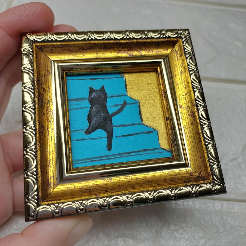 May include: A black cat painted on a blue background, climbing up a set of stairs. The painting is framed in a gold frame with a silver trim.