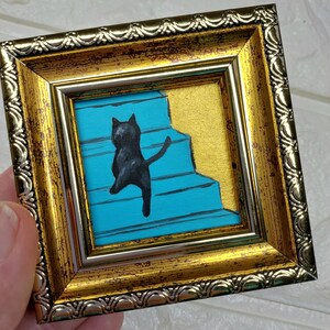 May include: A framed painting of a black cat walking up a blue staircase with a gold background.