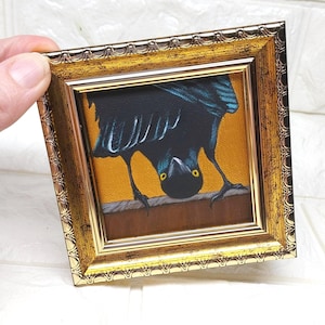 May include: A framed painting of a black bird with yellow eyes, set against a yellow background. The bird is upside down, perched on a brown ledge. The frame is gold with an ornate design.