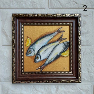 May include: A framed painting featuring three silver fish arranged diagonally on a textured gold background, accompanied by two yellow lemon wedges. The artwork is enclosed in a dark brown wooden frame with ornate gold detailing and a beaded inner edge.