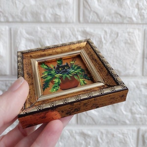 May include: A small framed painting of a black cat peeking out of a green potted plant. The frame is gold with a decorative border.