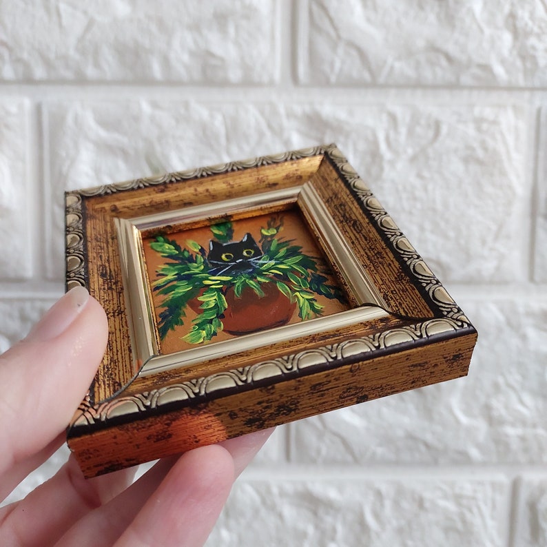May include: A small framed painting of a black cat peeking out of a green potted plant. The frame is gold with a decorative border.