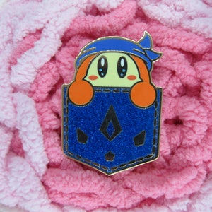 May include: A colorful enamel pin featuring a cartoon character with orange hair and a blue bandana peeking out of a blue glitter pocket. The pin has a gold outline and is set against a pink and white textured background.