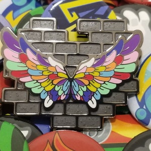 May include: A silver metal pin with a brick wall design and a colorful butterfly wing design. The wings are made up of multiple colors, including red, orange, yellow, green, blue, purple, and pink.